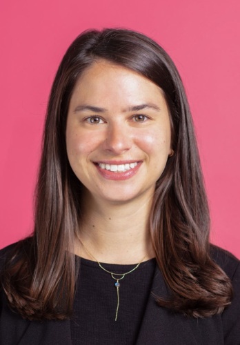Leah Greenberg, a woman with long brown hair, smiling in front of a pink background.